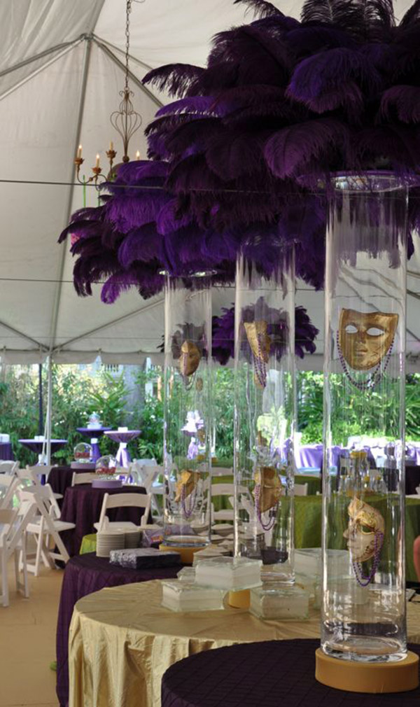Decorative table with tall glass vases filled with purple feathers and masks, set in a decorated event space with tables and chairs.