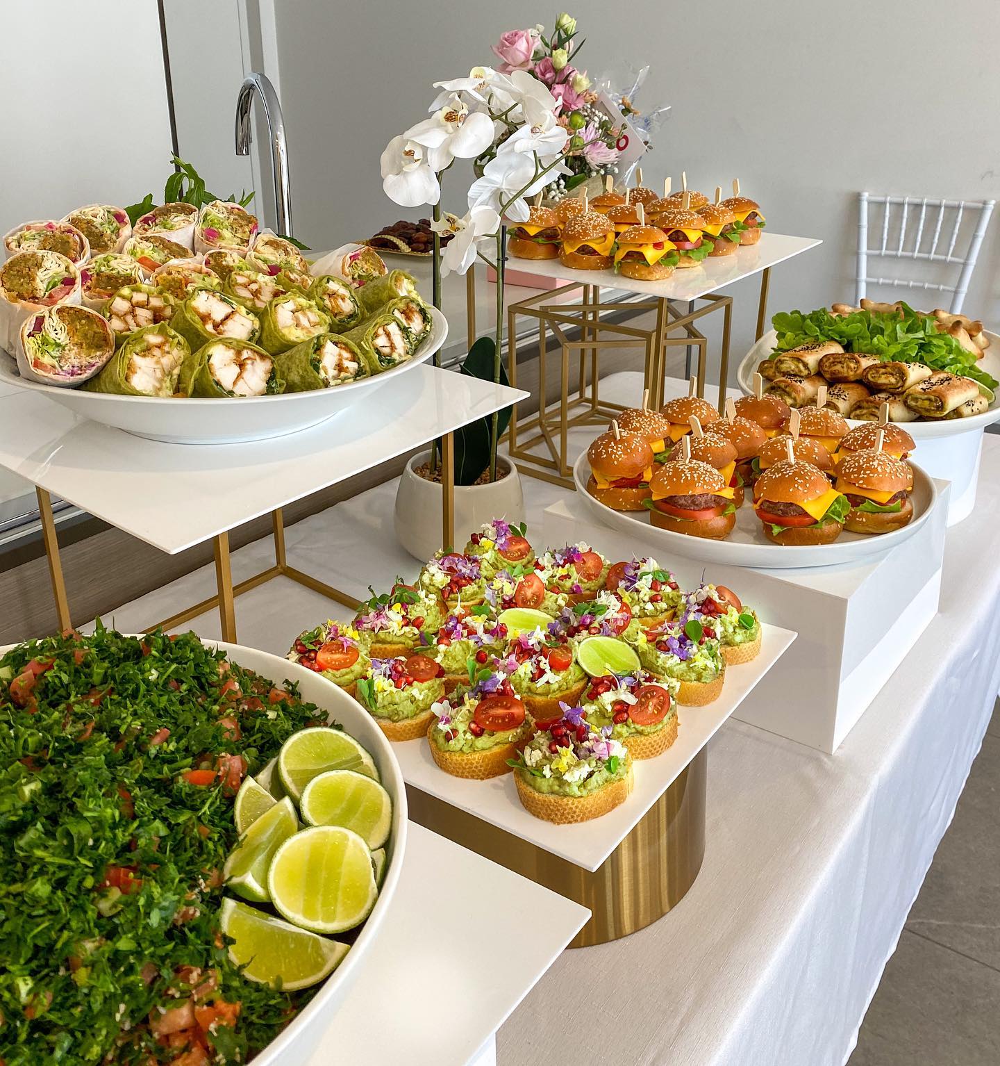 Table with assorted appetizers, cupcakes, and lime wedges, decorated with flowers, set in a bright room with white walls.