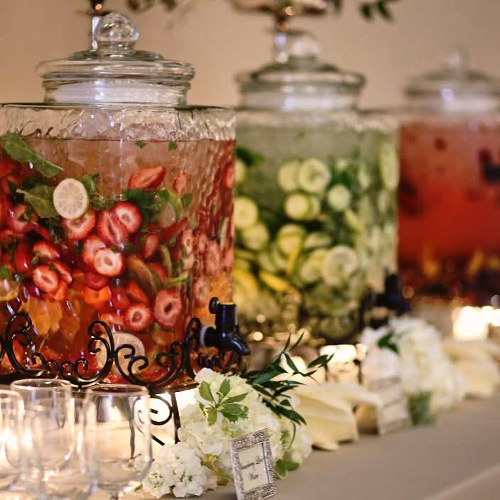 Three glass jars filled with colorful fruit-infused beverages, decorated with flowers and greenery, on a table with glasses and flowers.