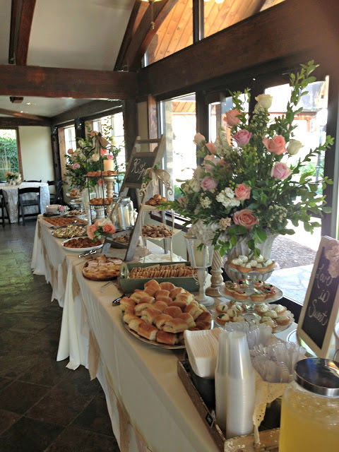 Table with floral arrangements and desserts in a bright room with large windows, wooden beams, and natural light.