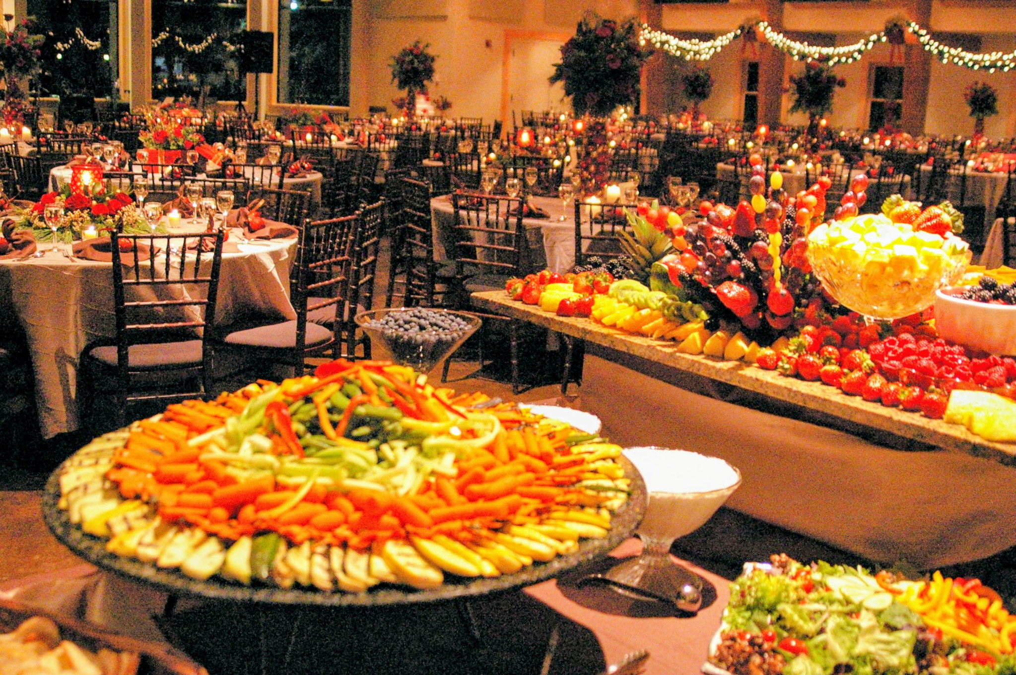 Banquet hall decorated with tables, chairs, and floral arrangements for celebration, with large fruit and vegetable displays in the foreground.