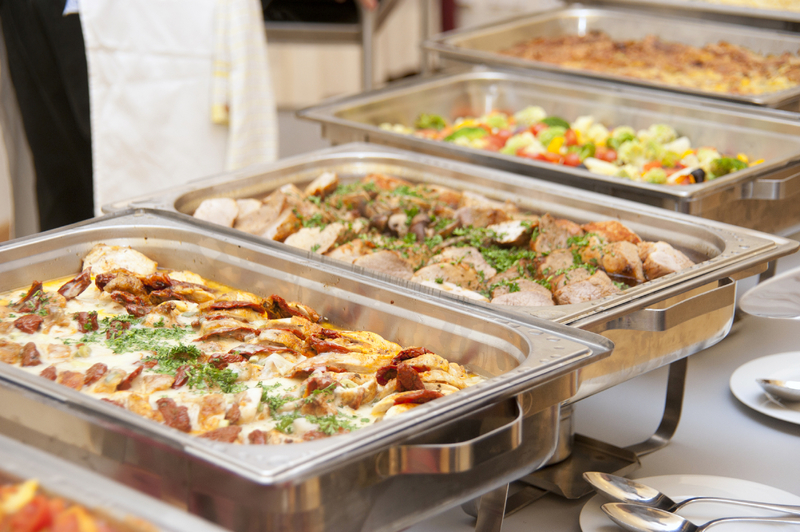 Three stainless steel chafing dishes with food, including pizza and cooked dishes, on a buffet table.