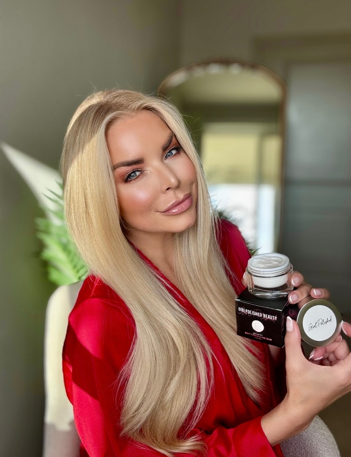 Woman with long blonde hair in a red top holding skincare products indoors, smiling slightly.