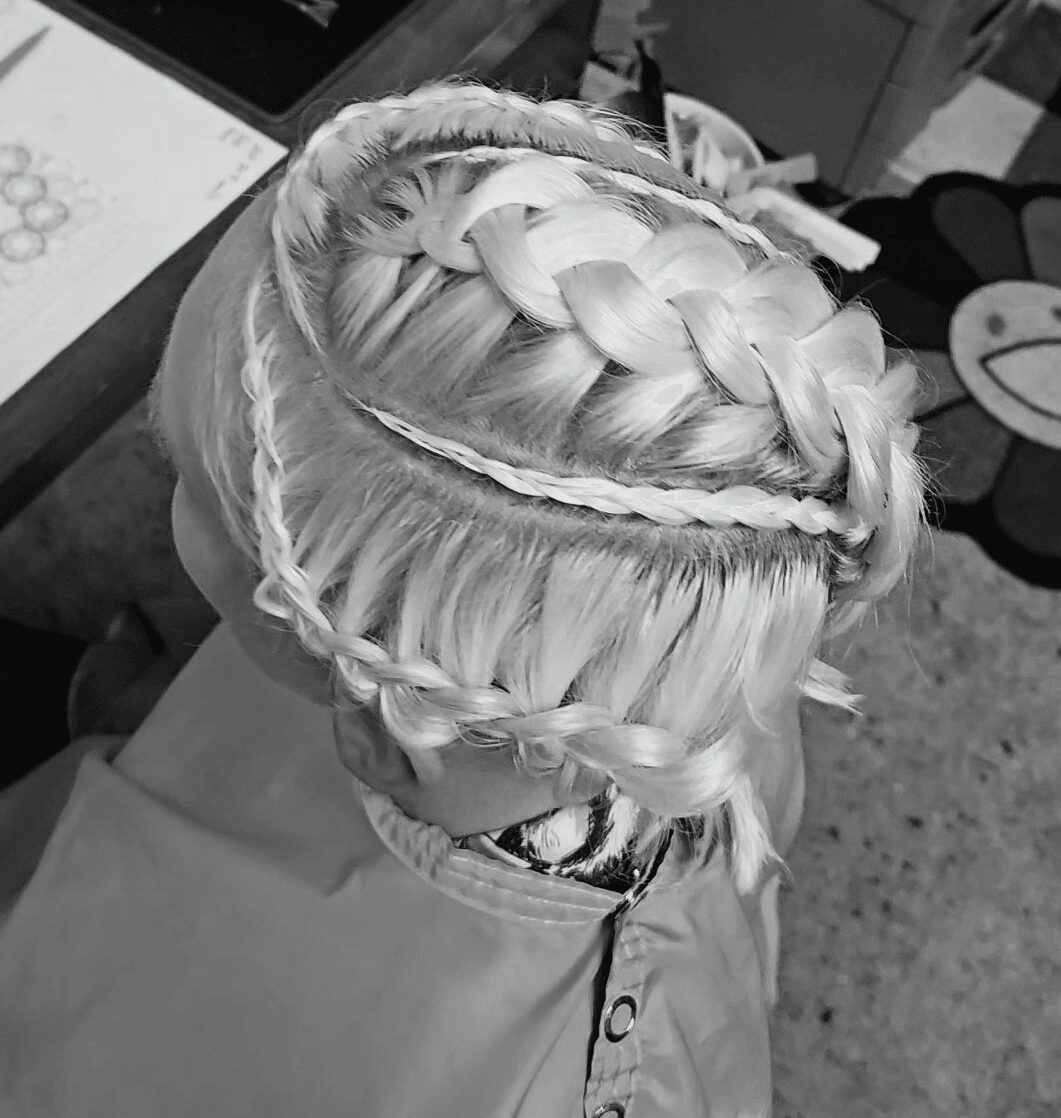Top view of a person with braided platinum blonde hair, wearing a cape, in a room with a smiley face pillow.