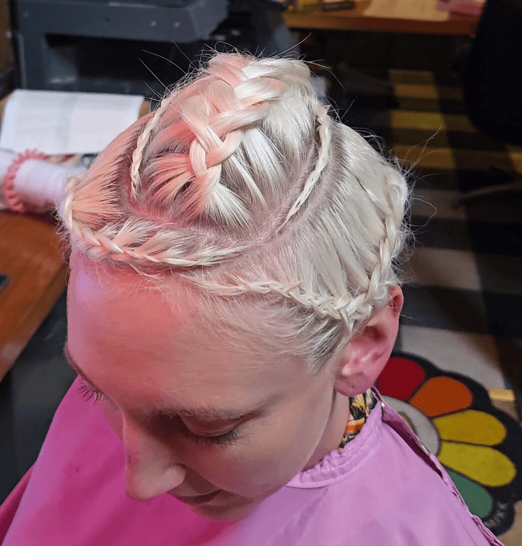 Young girl with platinum blonde hair styled in intricate braids, wearing a pink top, indoors with a colorful floor mat visible.