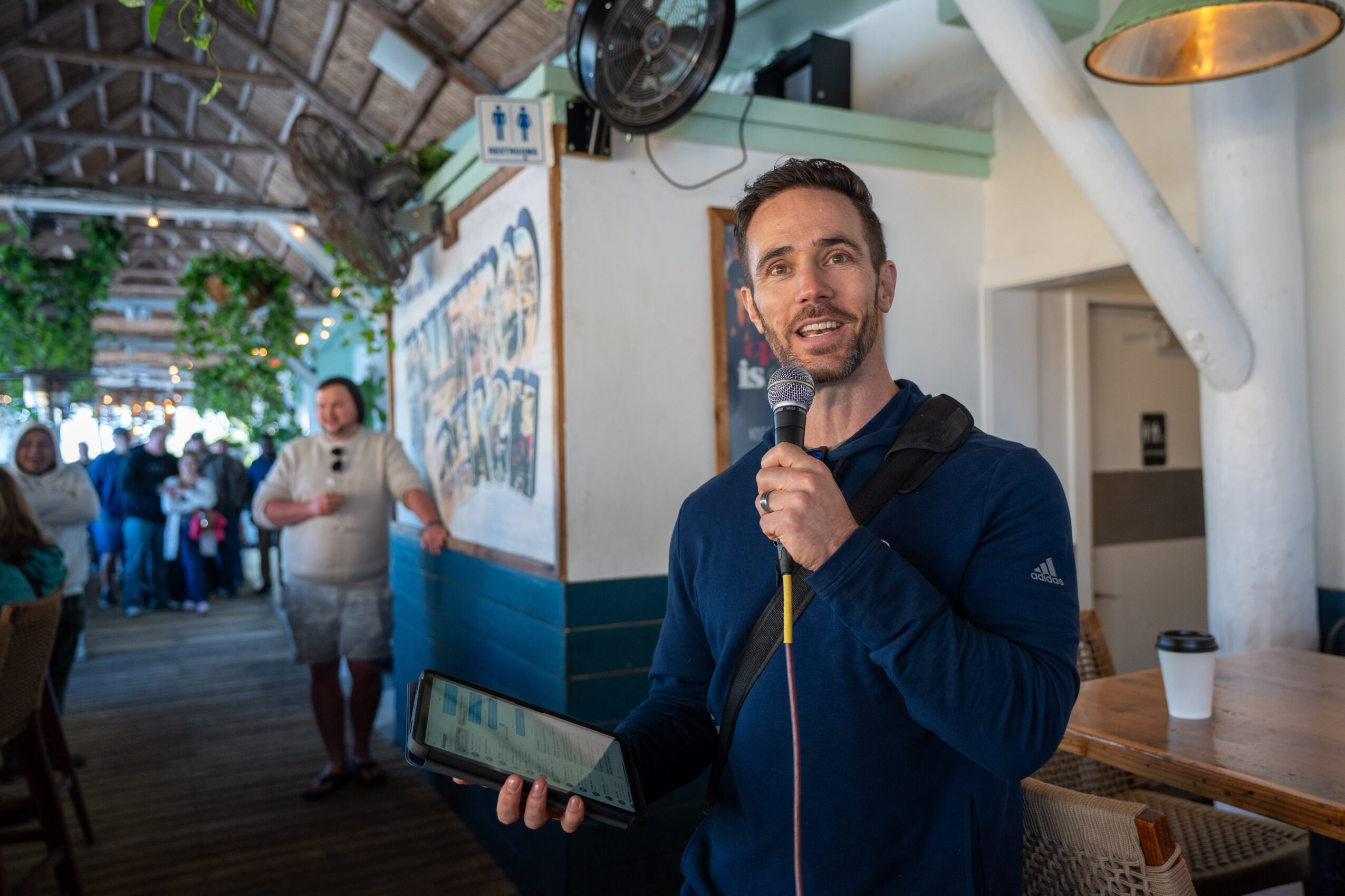 Man holding a microphone and tablet, smiling indoors with people and artwork in background.