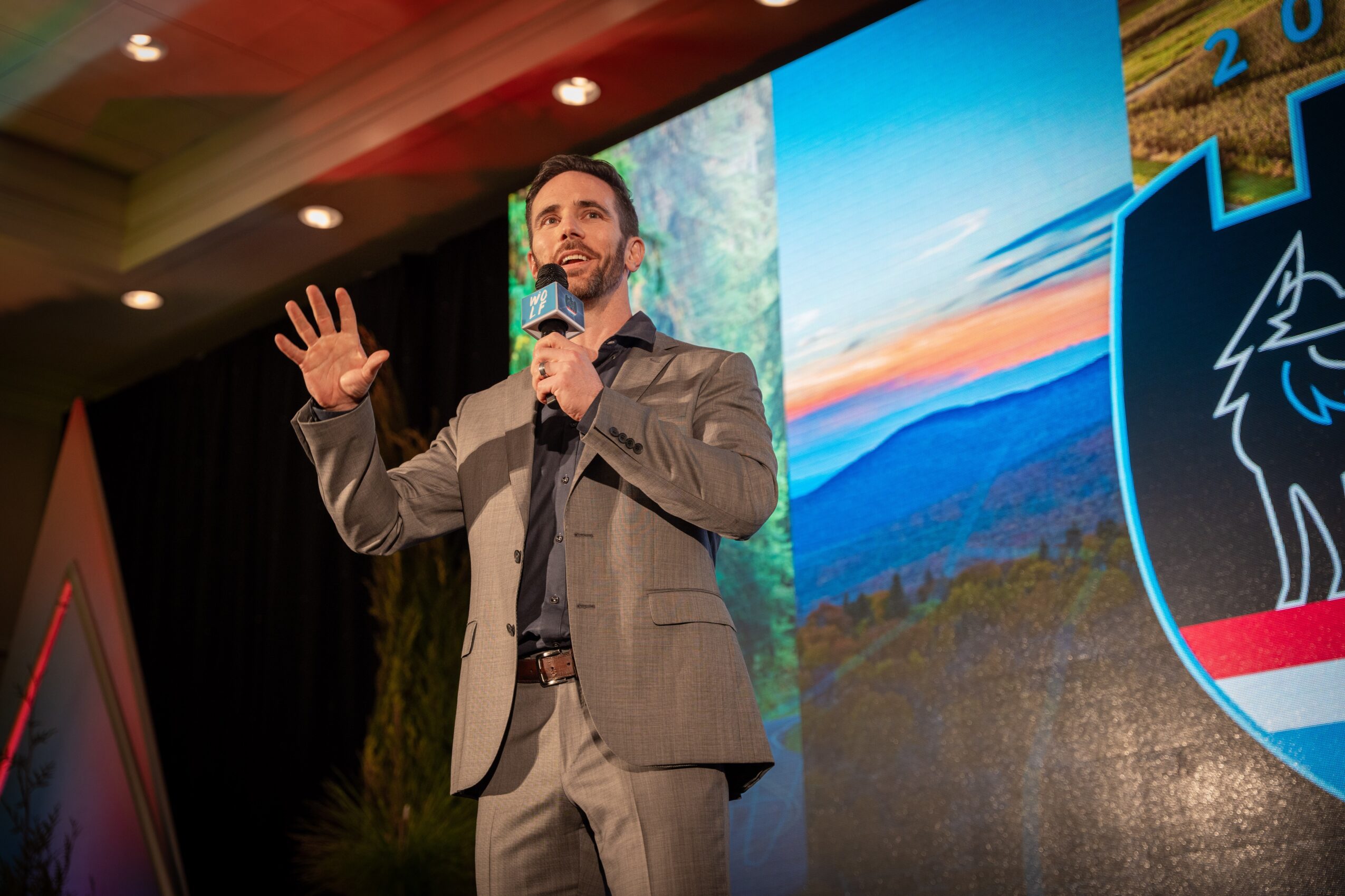 Man in a gray suit speaking into a microphone on stage with a large screen behind him showing a landscape and a logo.