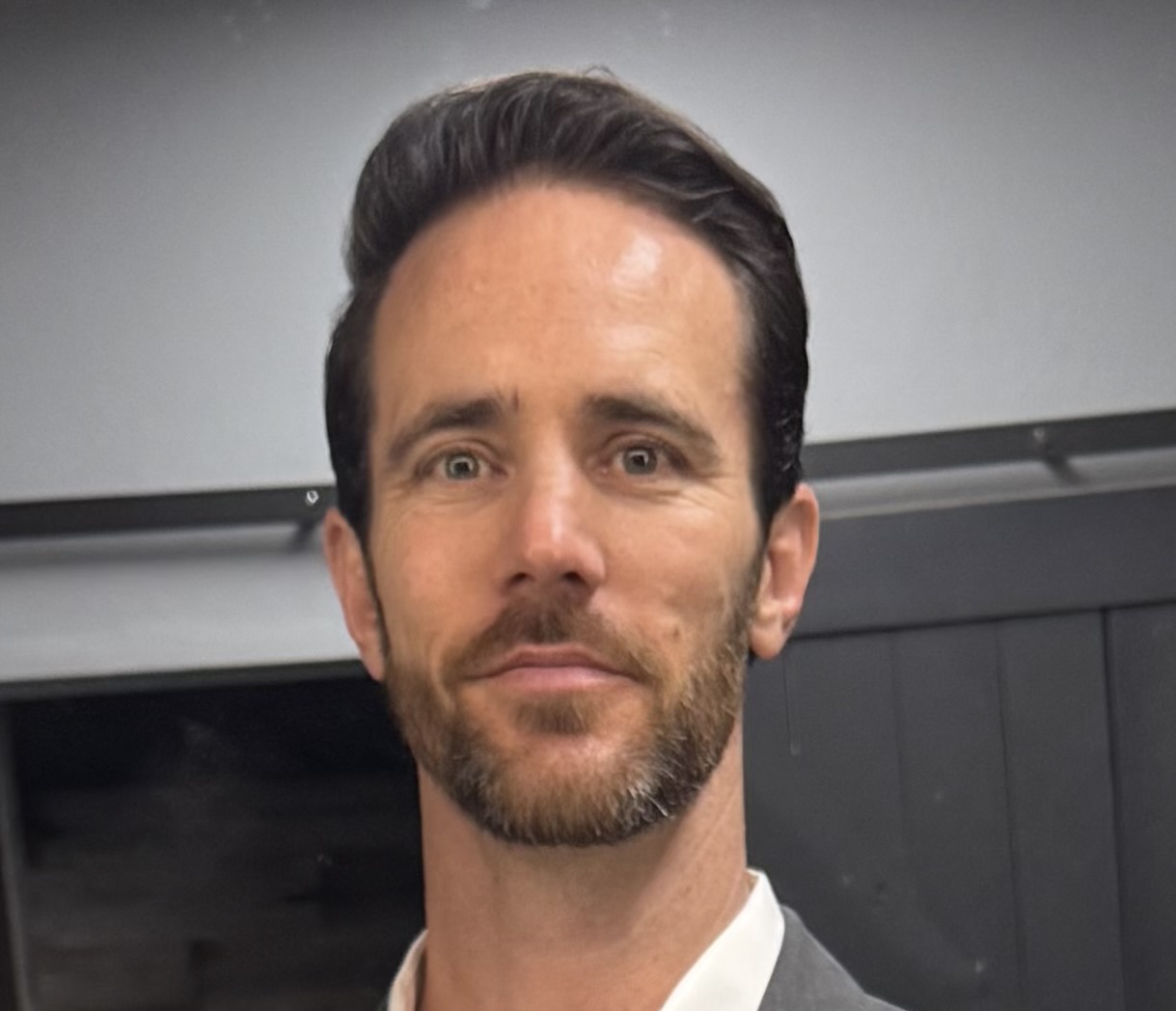 Man with dark hair and beard, wearing a suit, standing indoors with a neutral background.