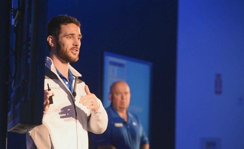 Two men on stage, one speaking and the other standing in the background, with a blue background and screen behind them.
