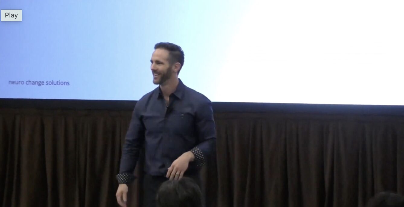 Man standing in front of a presentation screen, smiling, with a dark curtain below the screen.