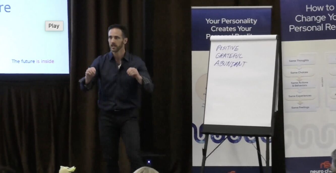 Man standing on stage next to a whiteboard with handwritten notes, in front of a presentation screen and banners.
