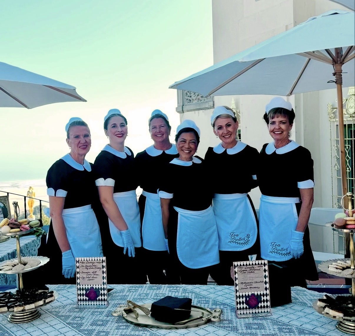 Six women dressed as waitresses in black uniforms with white aprons and hats, standing outdoors under umbrellas, smiling.