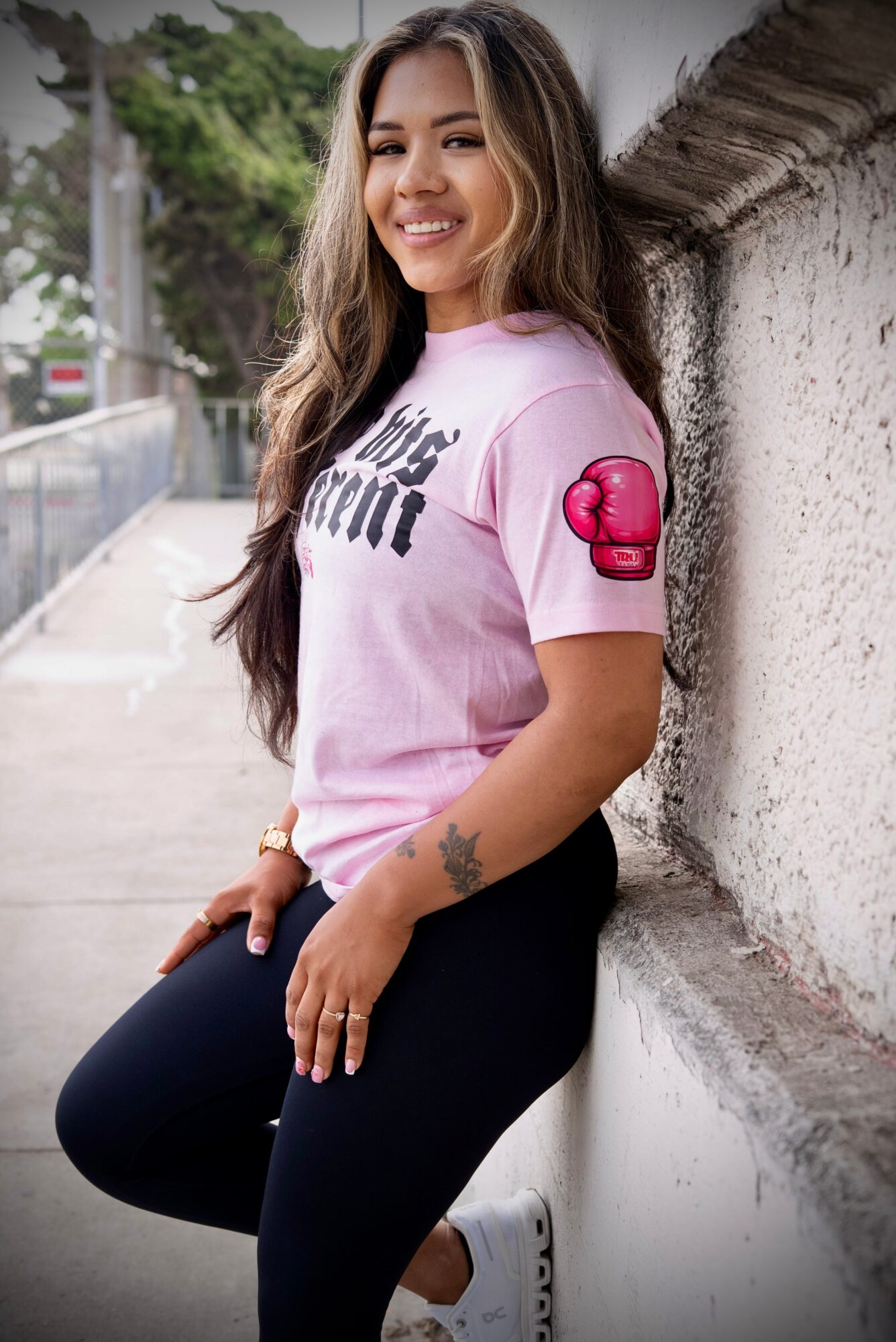 Young woman with long hair smiling, leaning against a wall outdoors, wearing a pink T-shirt and black pants.