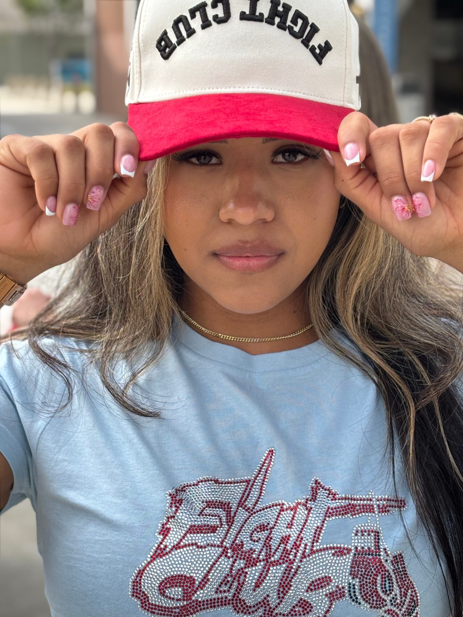 Young woman wearing a white and red cap, holding the brim, with long hair and a light blue shirt, outdoors.