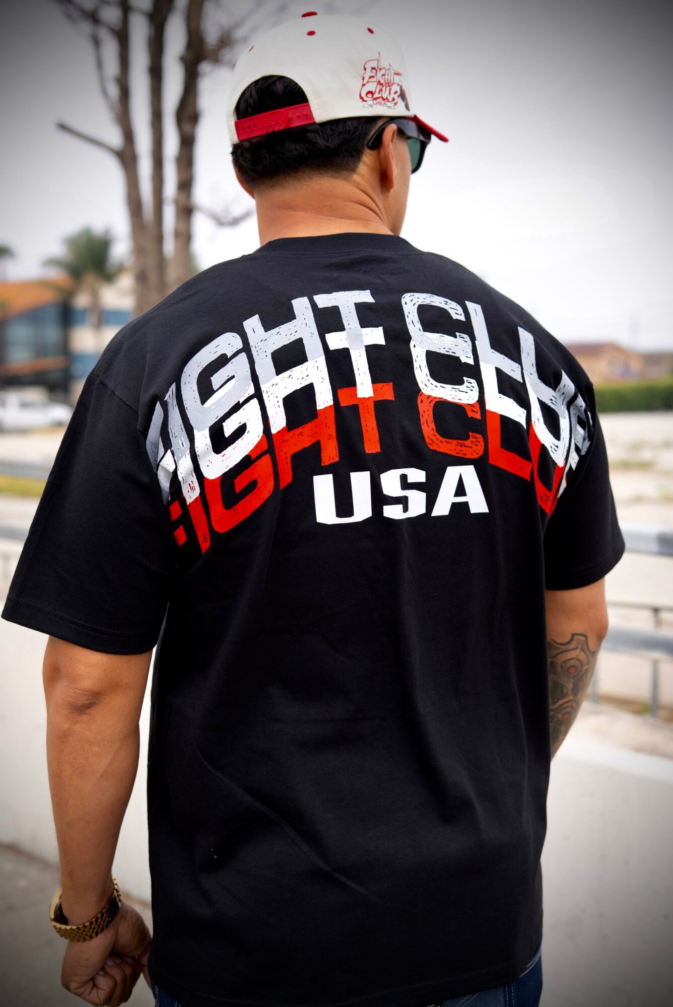 Person wearing a black T-shirt with bold white and red text, standing outdoors with trees and buildings in background.