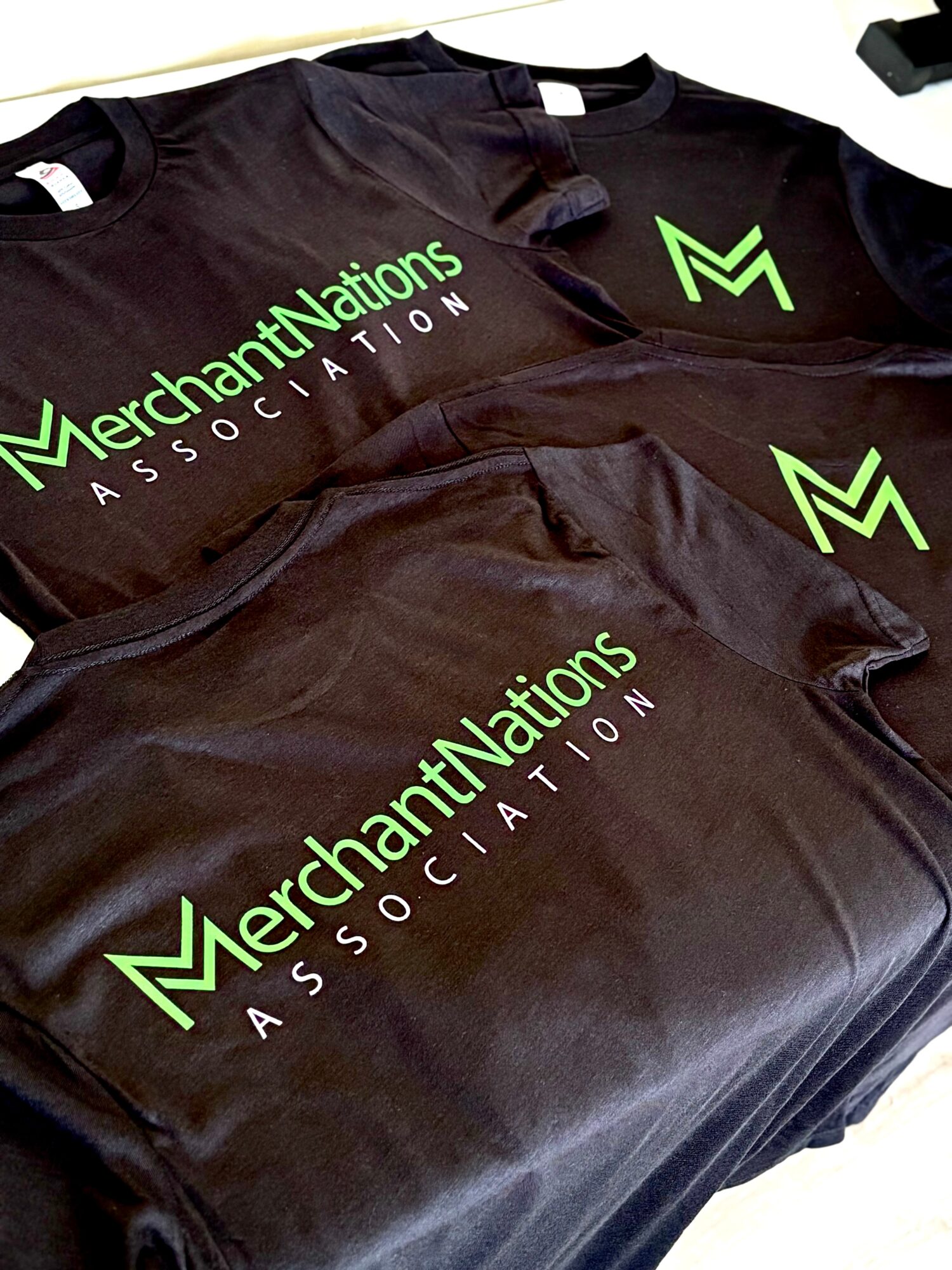 Three black T-shirts with green and white text and logo, laid flat. The text reads 'MerchantNations ASSOCIATION'.