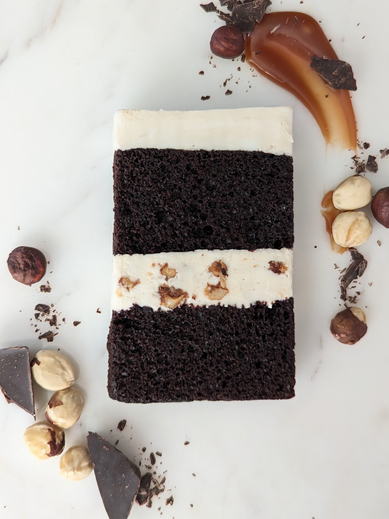 A layered chocolate cake with white frosting, garnished with nuts and chocolate pieces, on a white surface.
