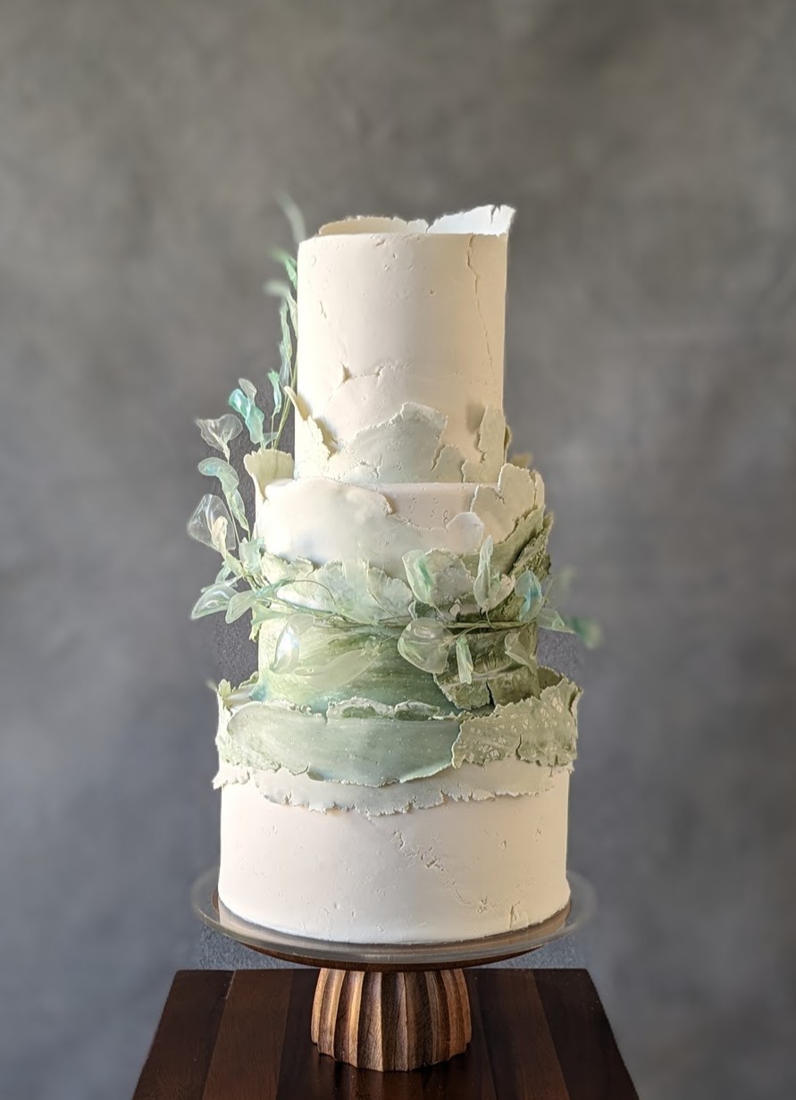 Tall, multi-layered white cake with green decorative elements on a wooden stand against a gray background.