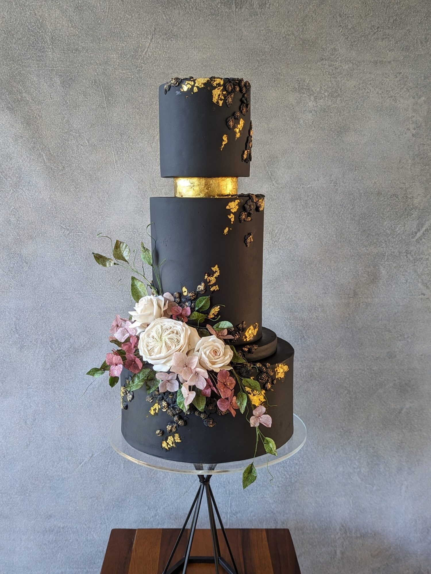 Three-tiered black cake with gold accents and floral decorations on a stand against a gray wall.