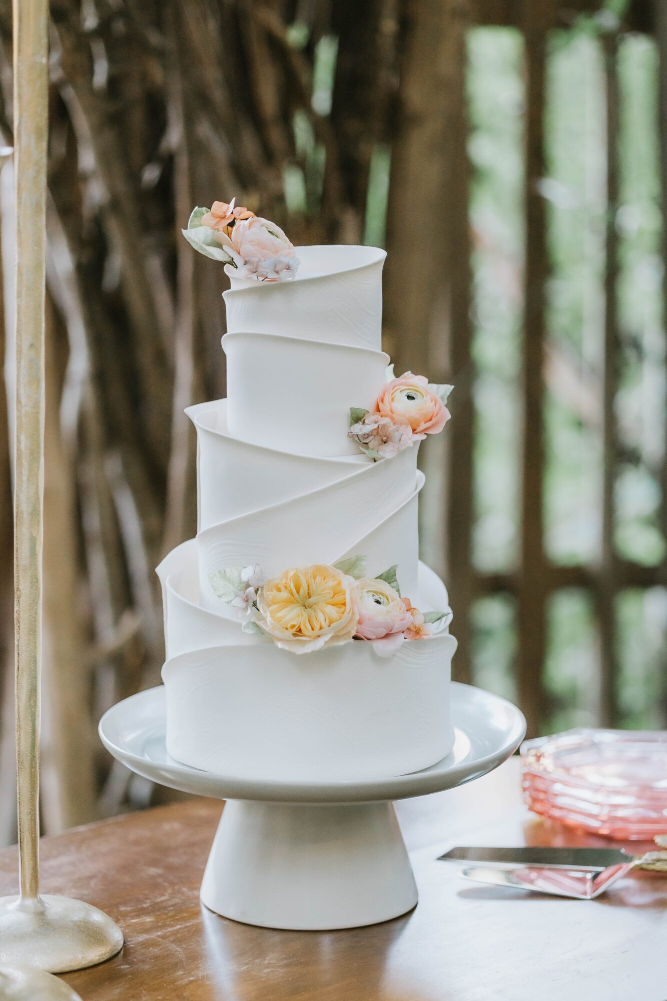 Tall, white, multi-tiered cake with flowers on top, displayed on a white stand outdoors with trees in background.