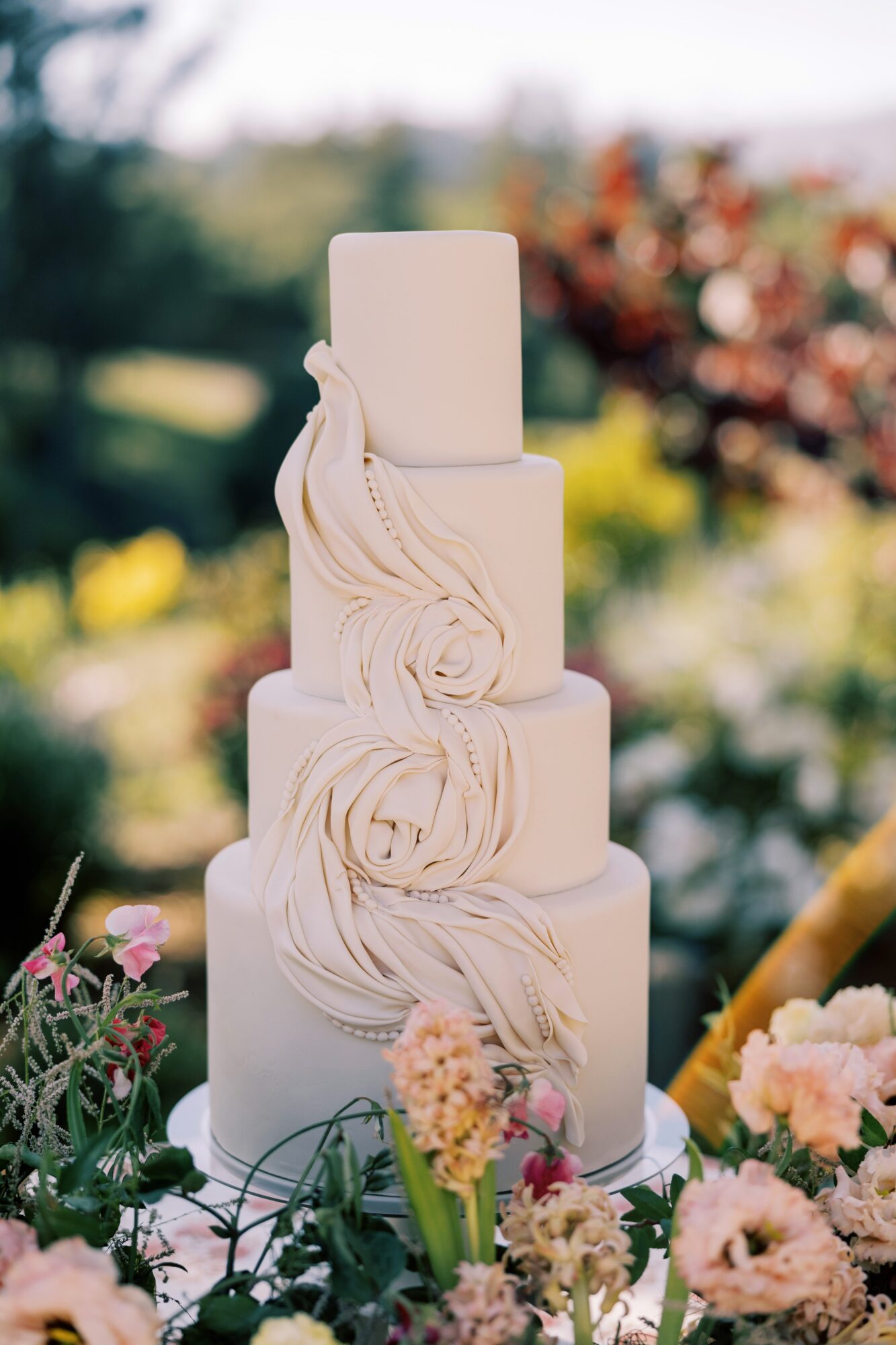 Tall, white, four-tiered cake with floral decorations, set outdoors with greenery and flowers in background.