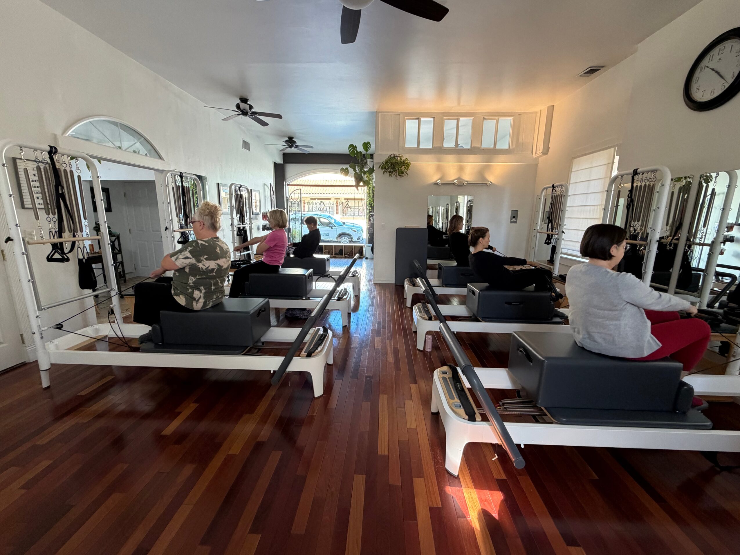 People exercising on Pilates reformers in a bright studio with large windows and wooden floors.