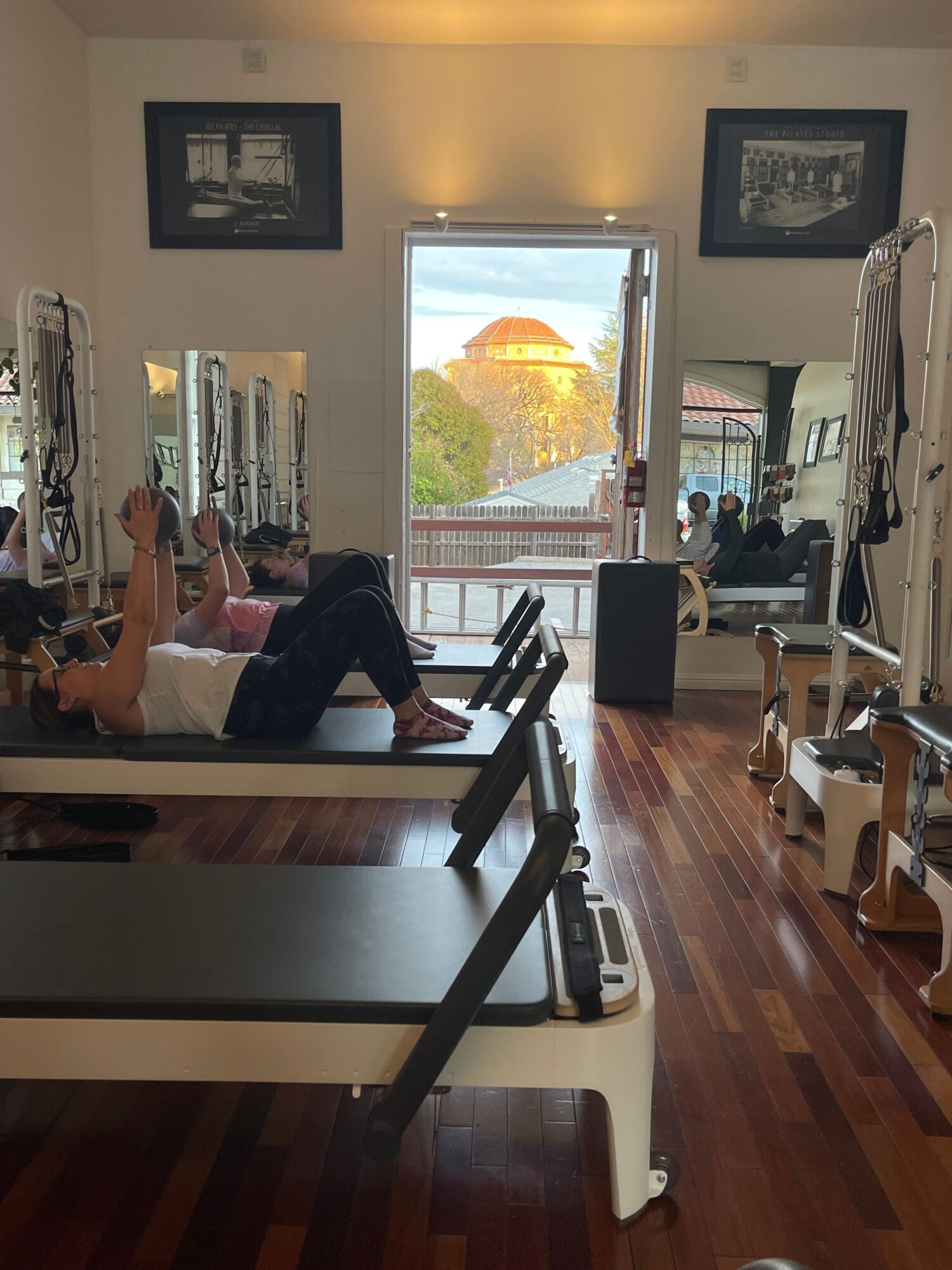 Person sitting on a bed in a room with wooden floors, window showing trees and sky, exercise equipment nearby.