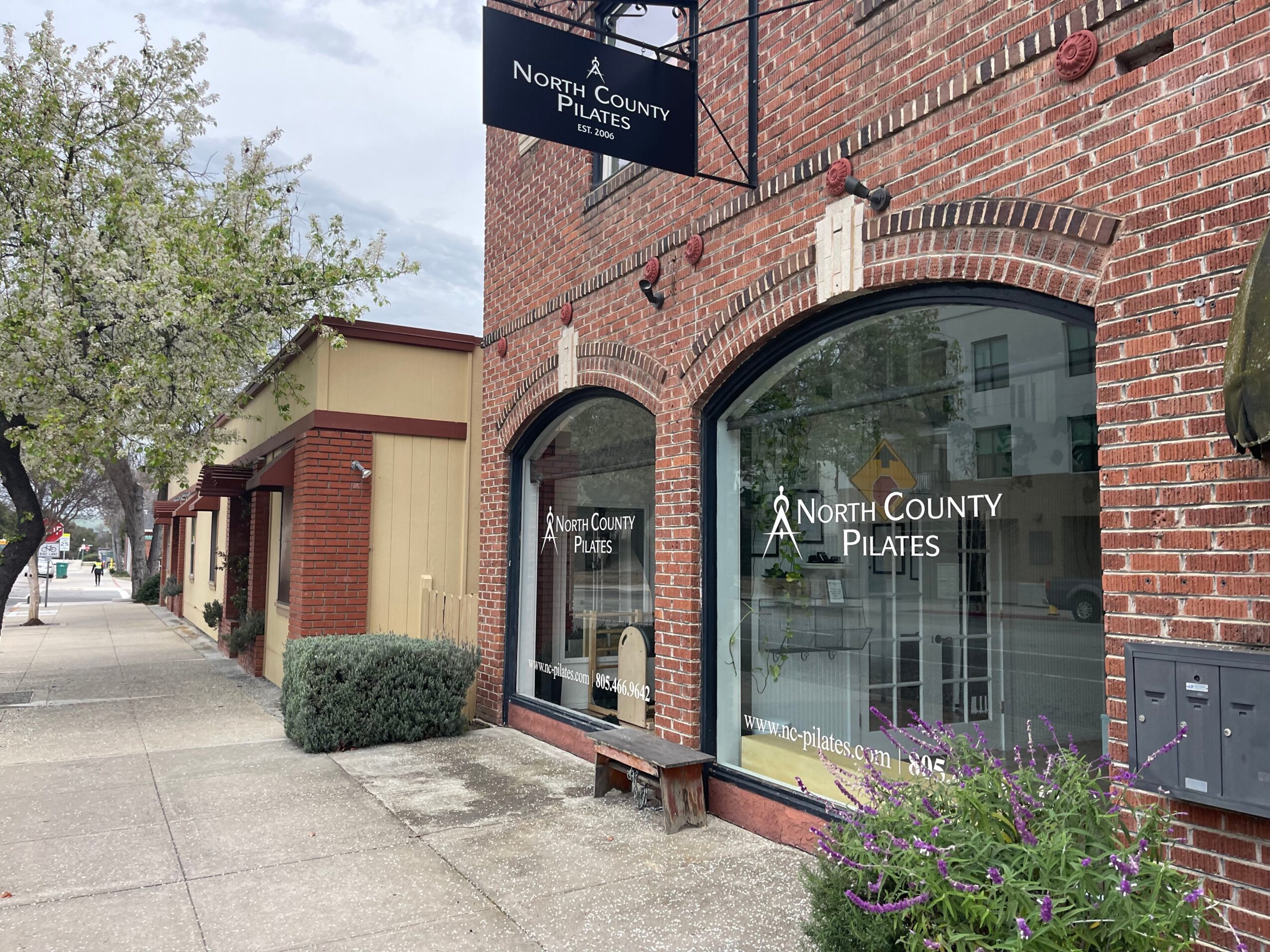 Brick building with large glass window and door, sign reading 'North County Pilates,' sidewalk with trees and bushes, cloudy sky.