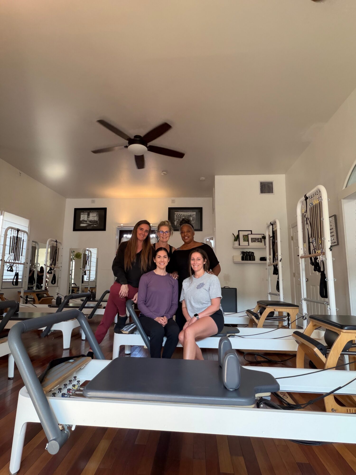 Five people in a room with exercise equipment, some sitting and some standing, with a ceiling fan overhead.