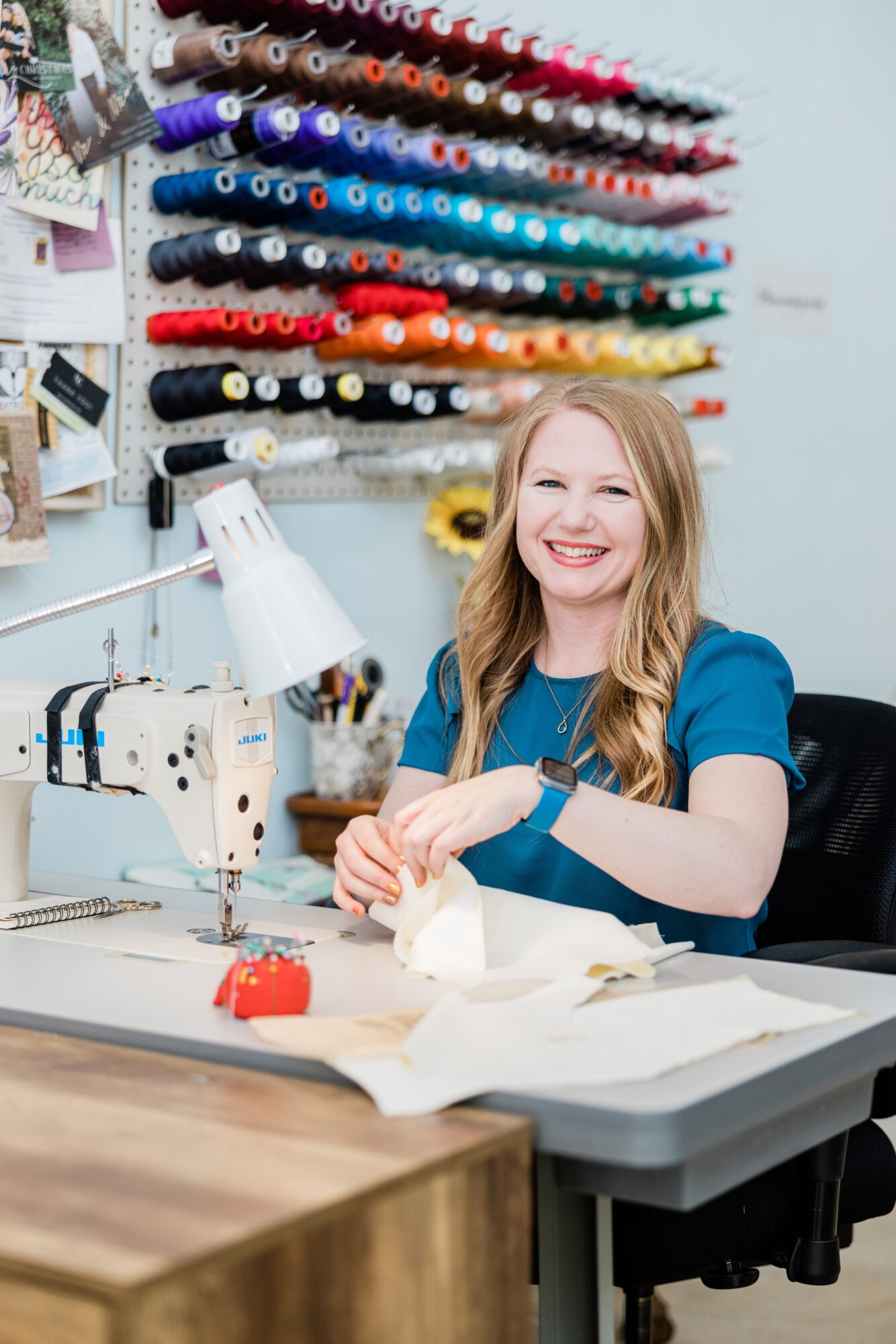 Young woman with long blonde hair smiling at sewing machine in fabric store, colorful thread spools on wall behind her.