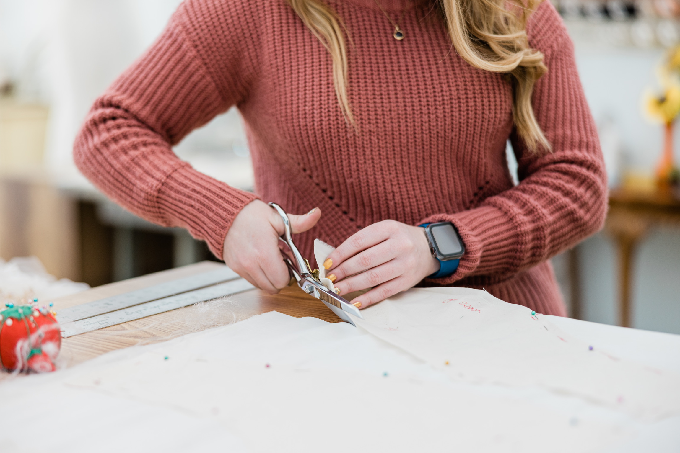 Person in a red sweater cutting fabric with scissors on a table, with a watch on their left wrist.