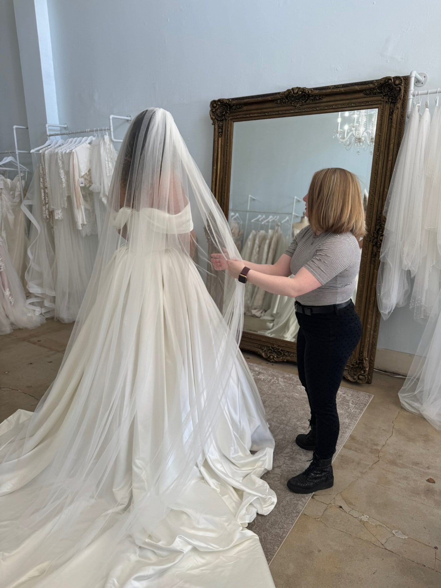 Person adjusting a wedding dress on a mannequin in a room with a large mirror and white curtains