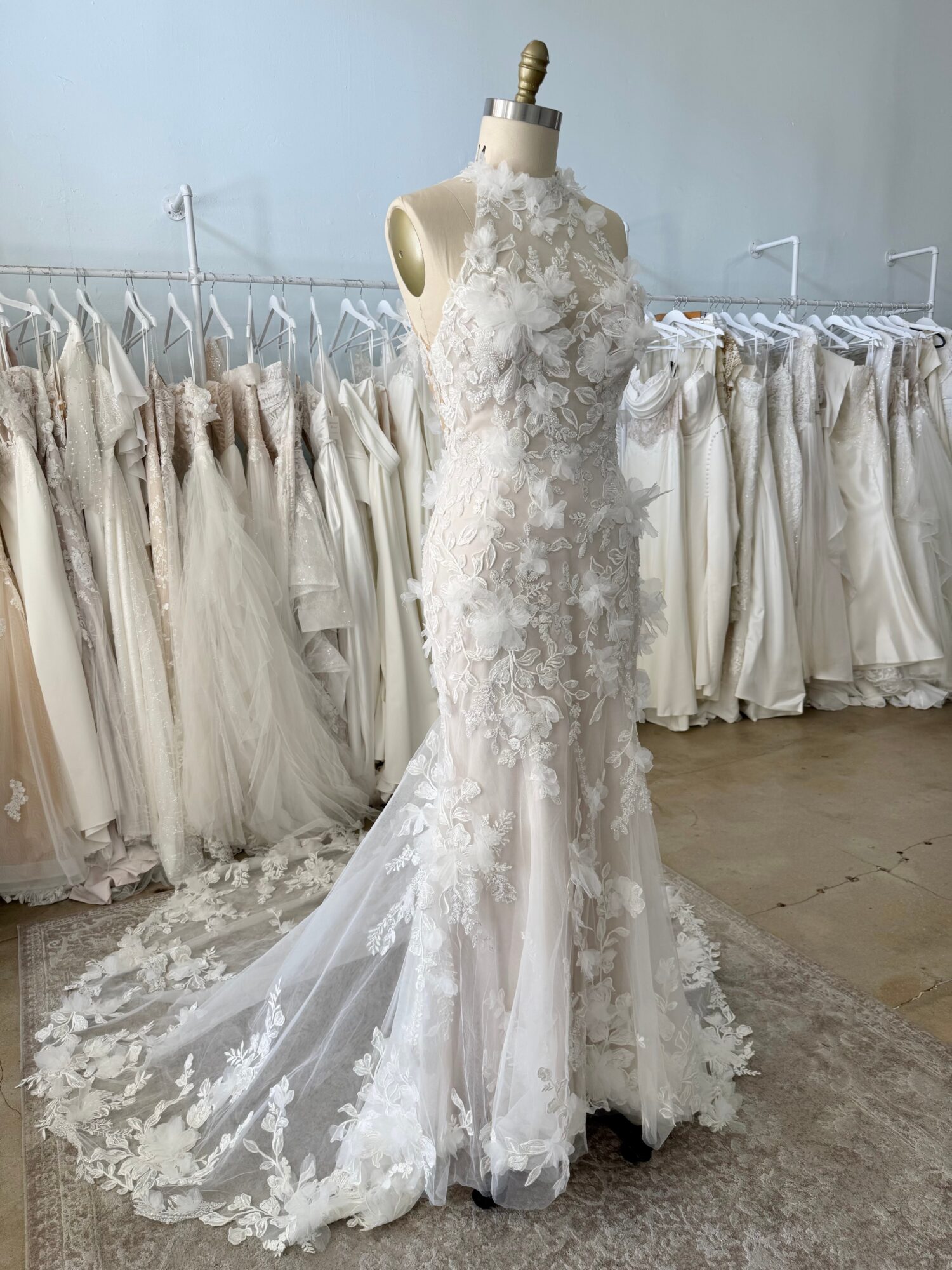 White lace wedding dress on mannequin with a train, surrounded by hanging wedding gowns in a bridal shop.