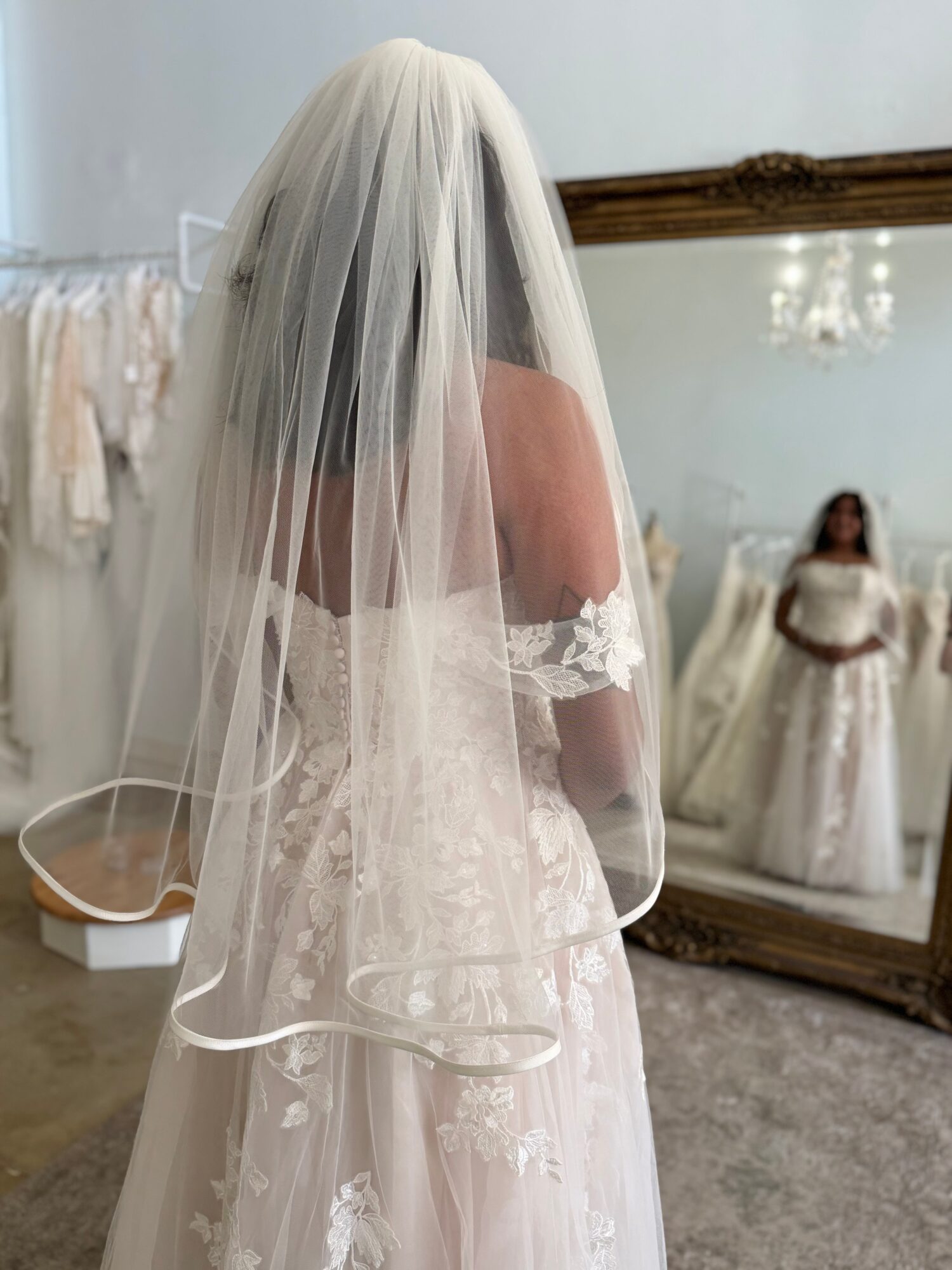 Bride with long veil and off-shoulder wedding dress looking at mirror, back view, in a room with chandelier.
