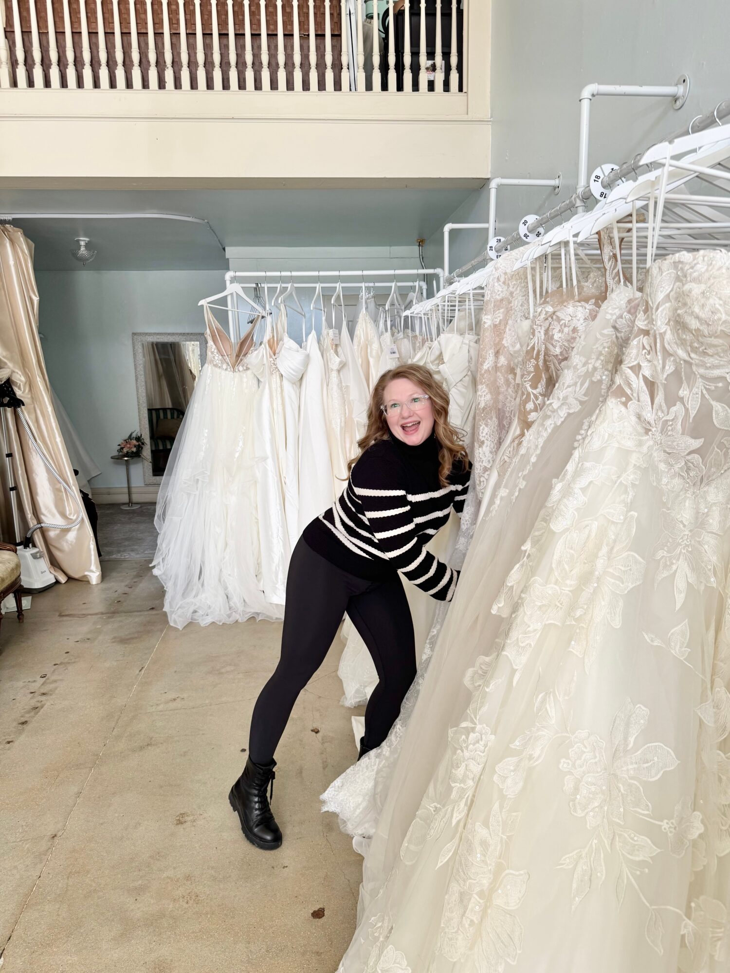 Young woman with long hair smiling and trying on wedding dresses in a bridal shop, surrounded by hanging gowns.