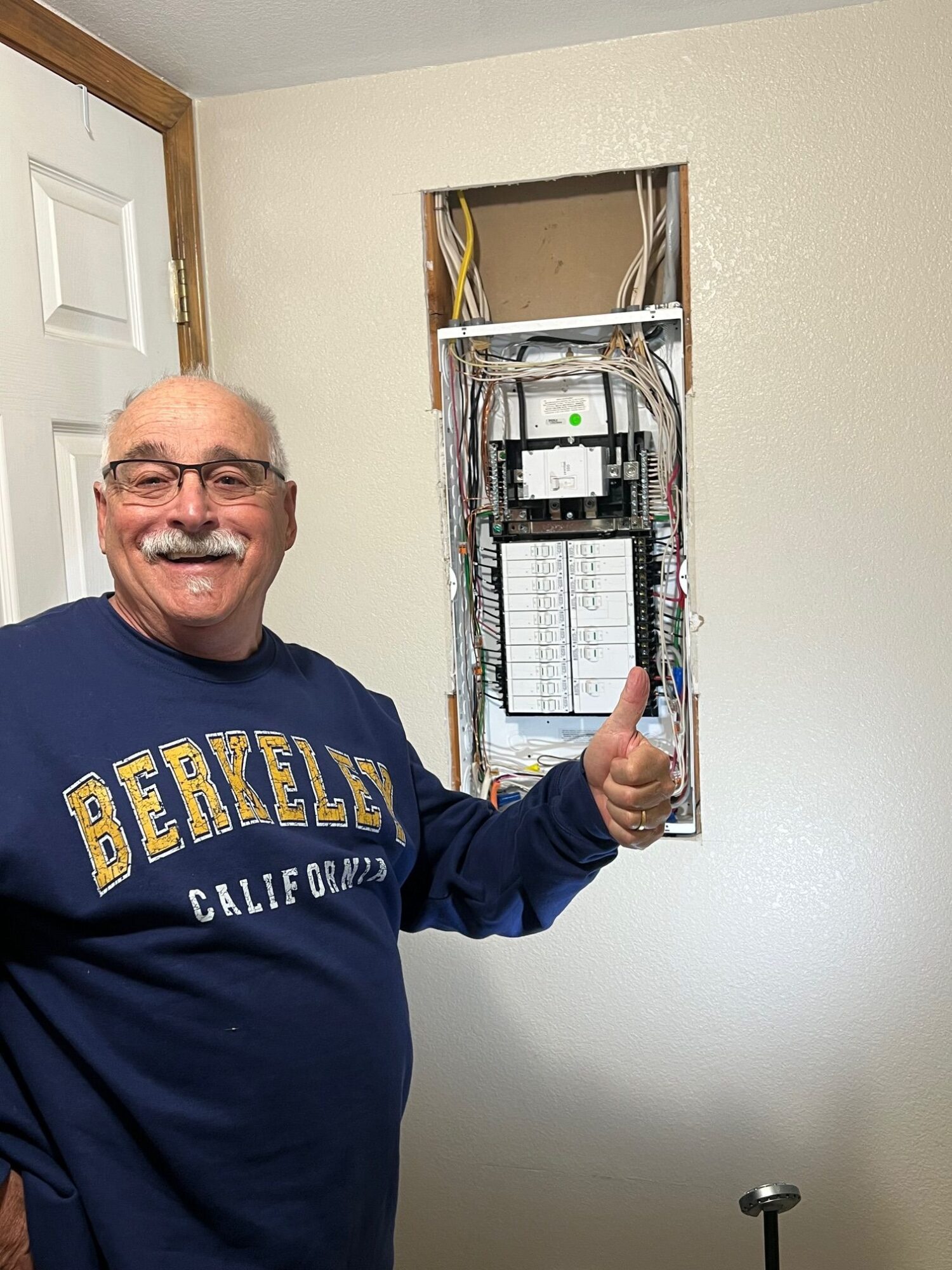 Man with glasses and a beard smiling, wearing a blue sweatshirt, standing next to an open electrical panel in a wall.