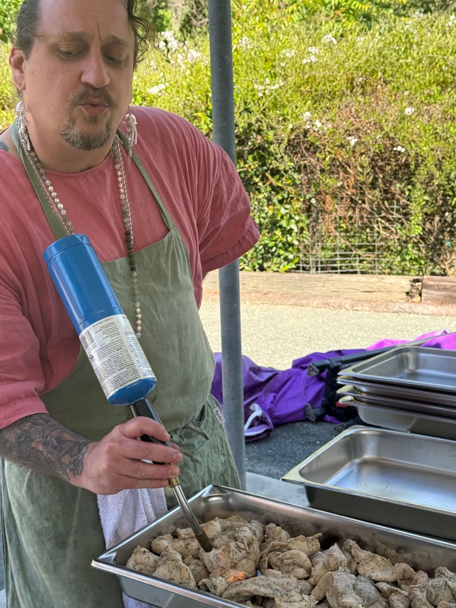 Man with tattoos and a beard, wearing a red shirt and green apron, holds a blue bottle near a grill with rocks.