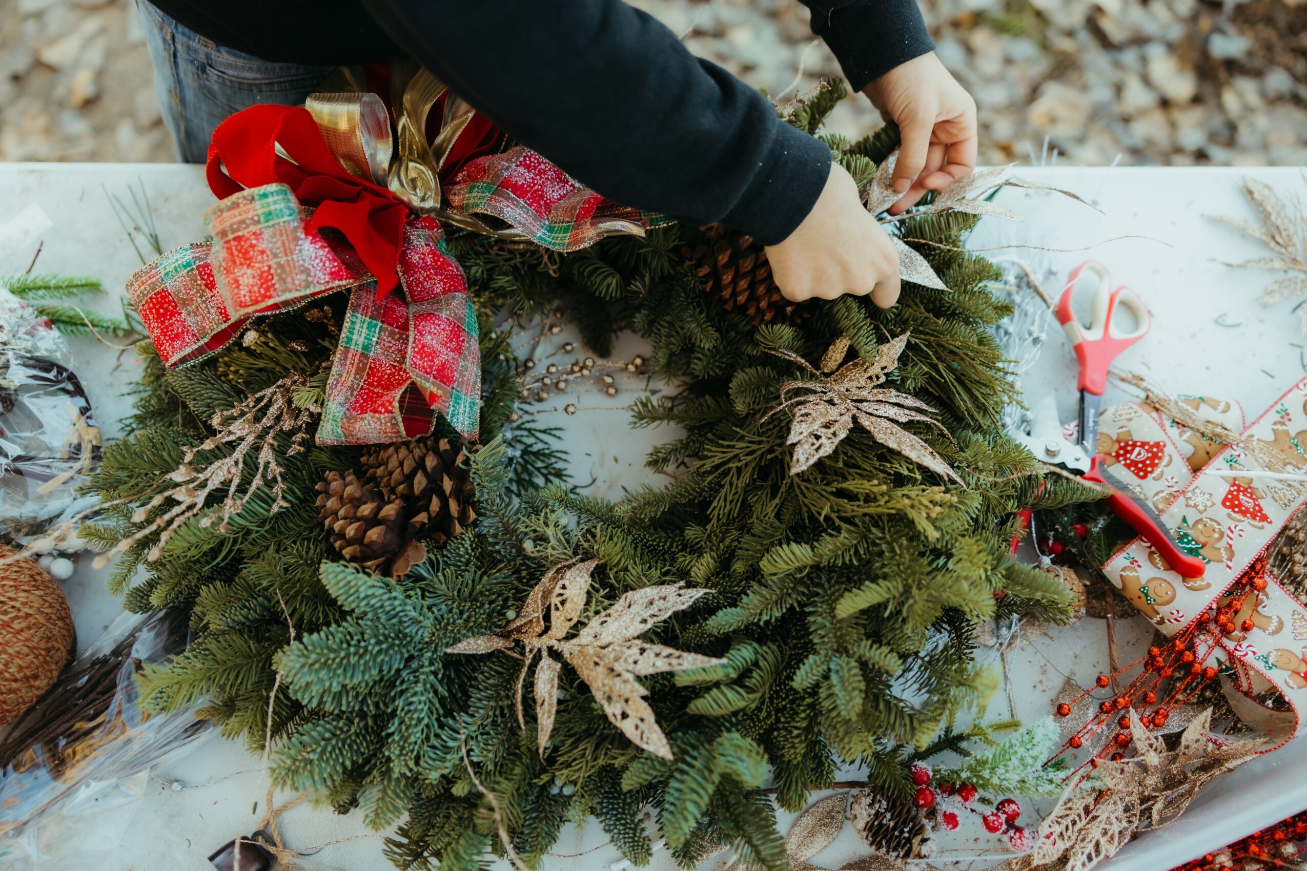 Person decorating a Christmas wreath with pinecones, ribbons, and foliage on a table outdoors.