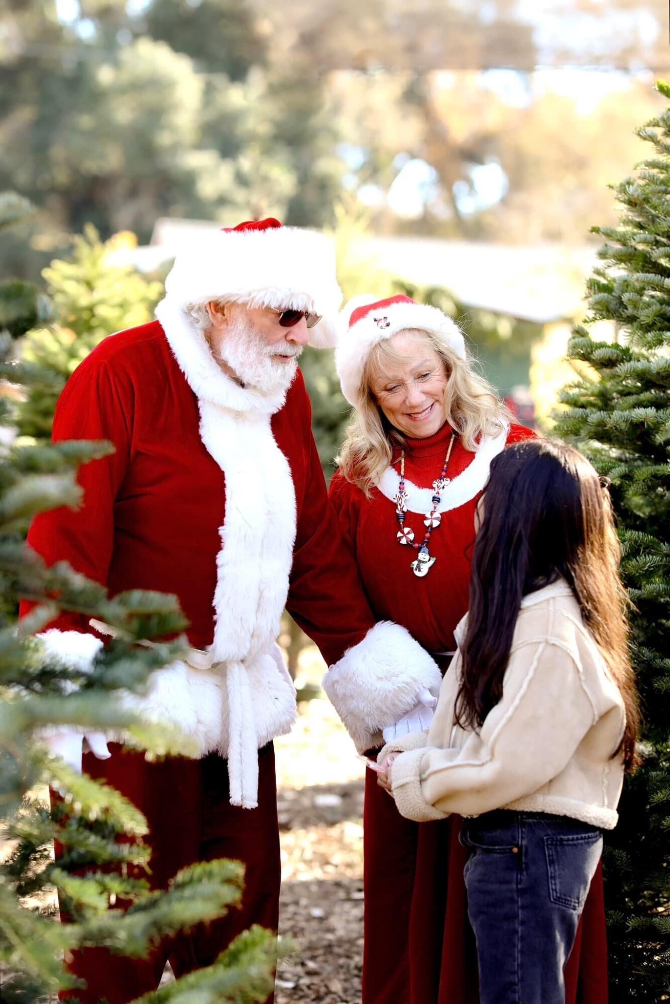 Two adults dressed as Santa and Mrs. Claus talking to a girl in a beige coat outdoors during Christmas, surrounded by trees.