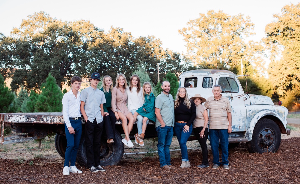 Group of nine people standing and sitting on a vintage truck outdoors with trees and sky in background.