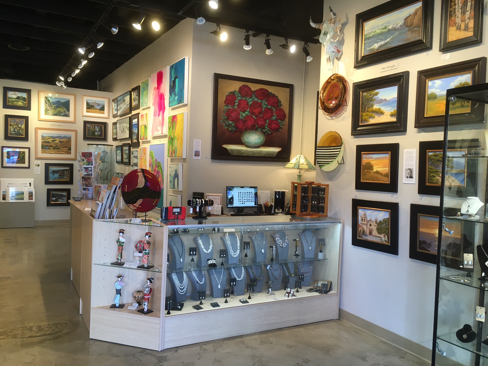 Art gallery with paintings on walls, a display counter with jewelry, and various framed artworks.