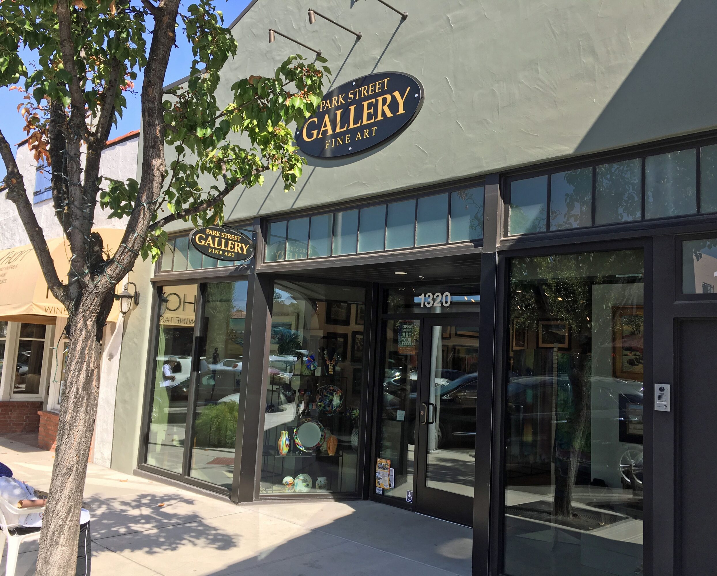 Gallery art gallery storefront with large glass windows and a sign above the entrance, tree in foreground, sunny day.