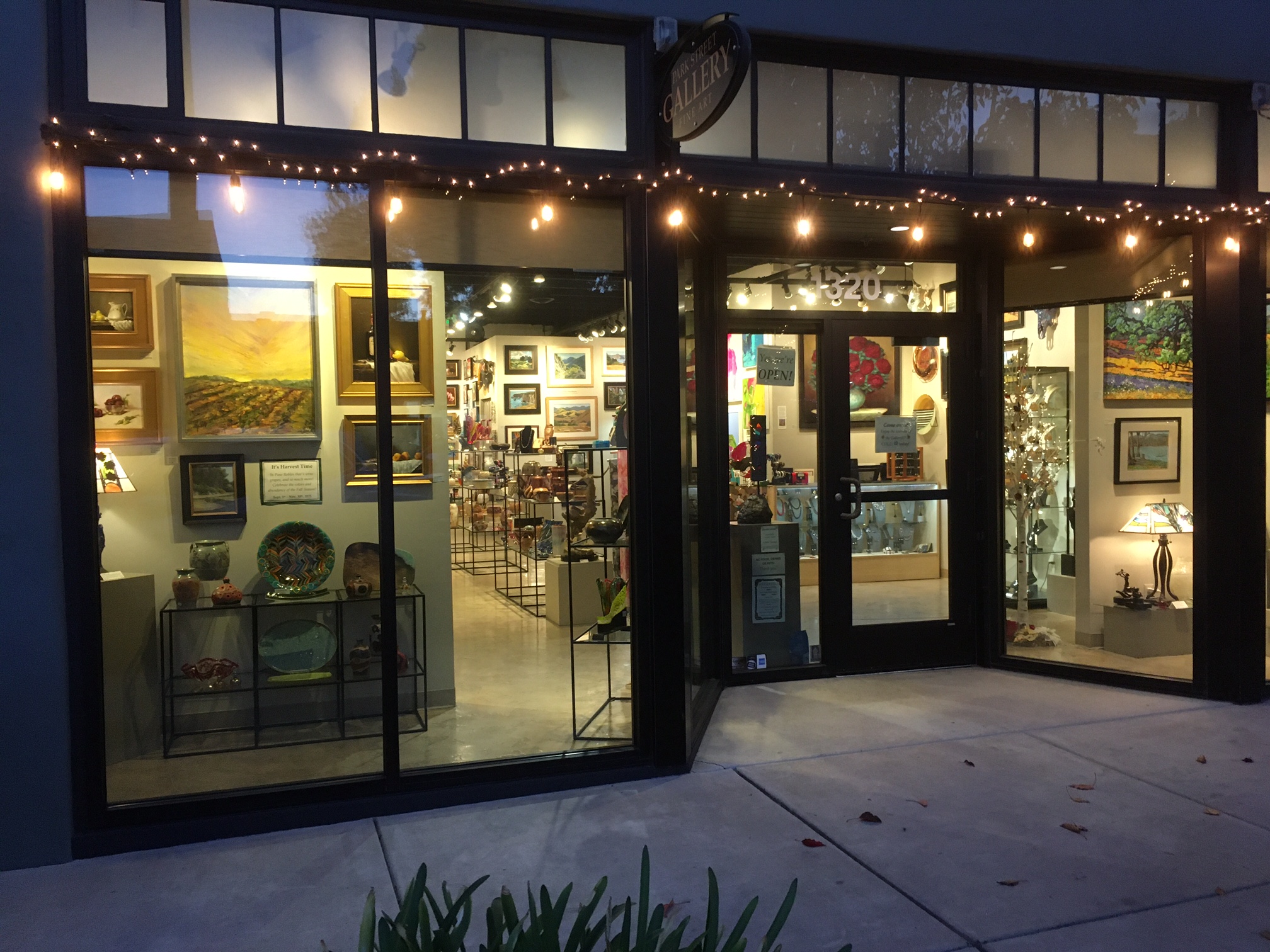 View of an art gallery with large glass windows, displaying framed artwork and interior lighting, outside sidewalk visible.