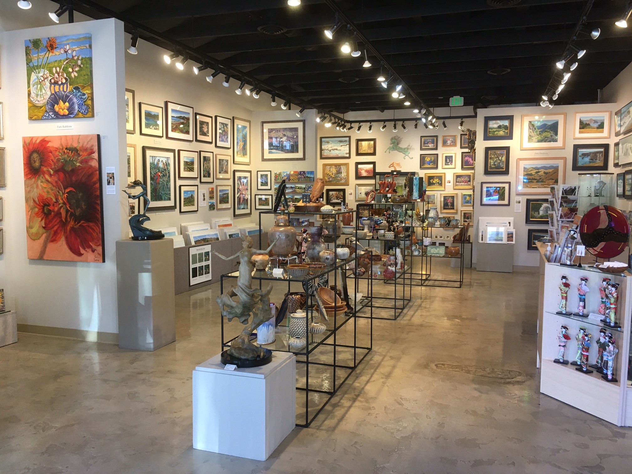 Art gallery with framed paintings on walls, sculptures on pedestals, and display cases, with a polished concrete floor and black ceiling.