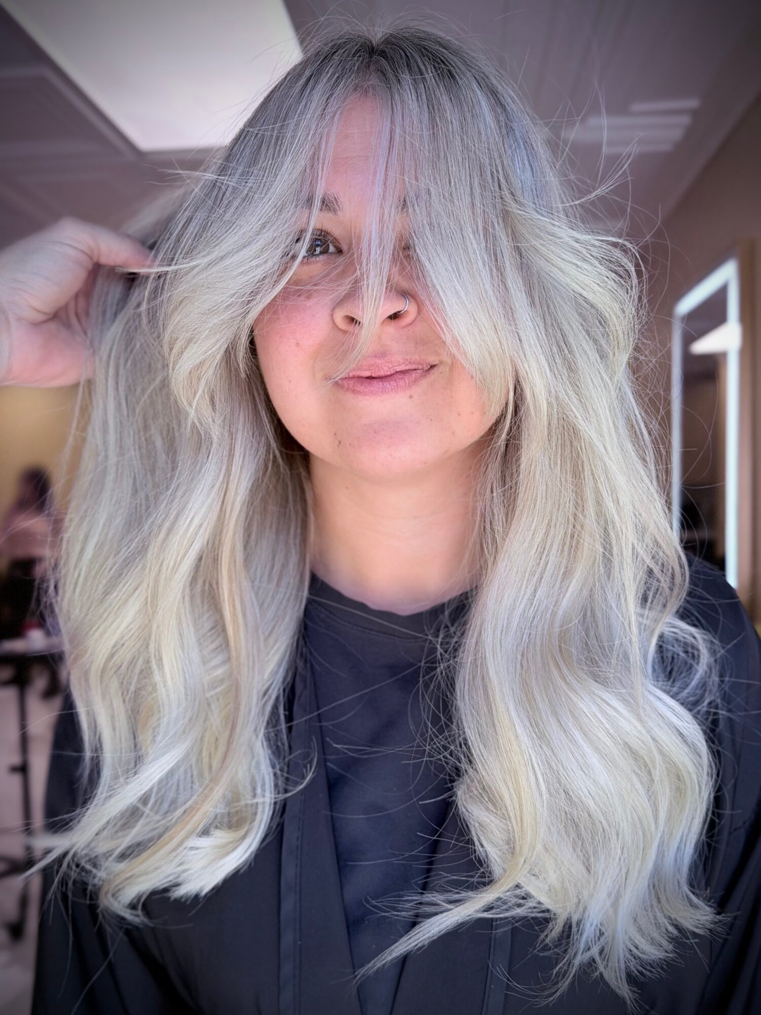 Woman with long, wavy, platinum blonde hair smiling, indoors with natural light, wearing a dark top.
