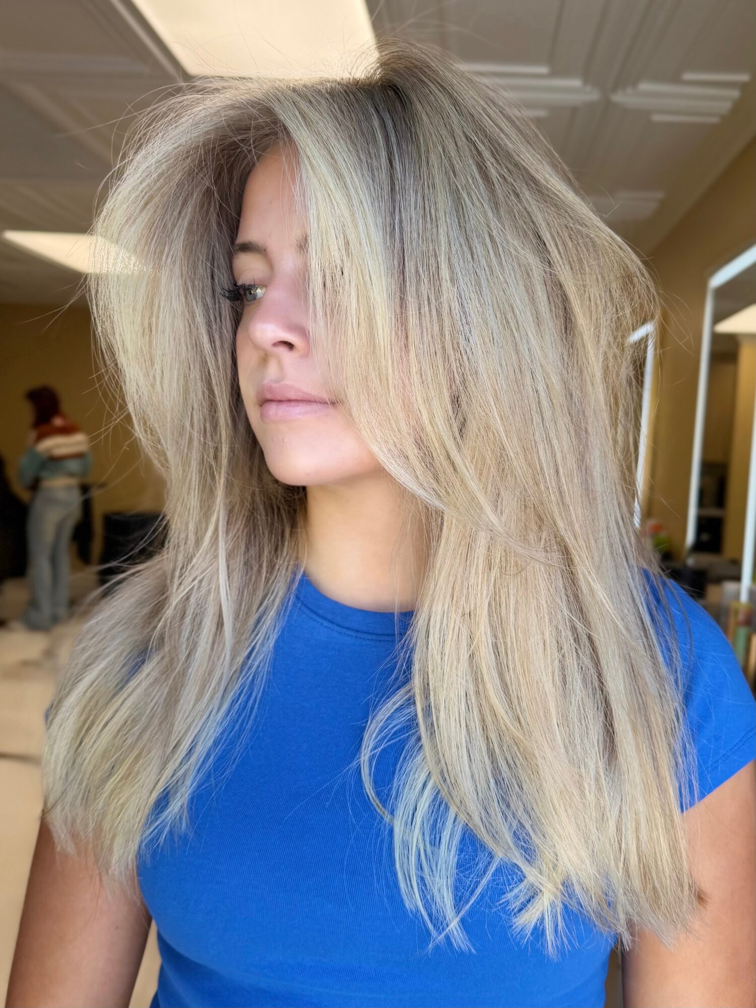 Woman with long, voluminous blonde hair wearing a blue shirt, indoors with mirrors and people in background.