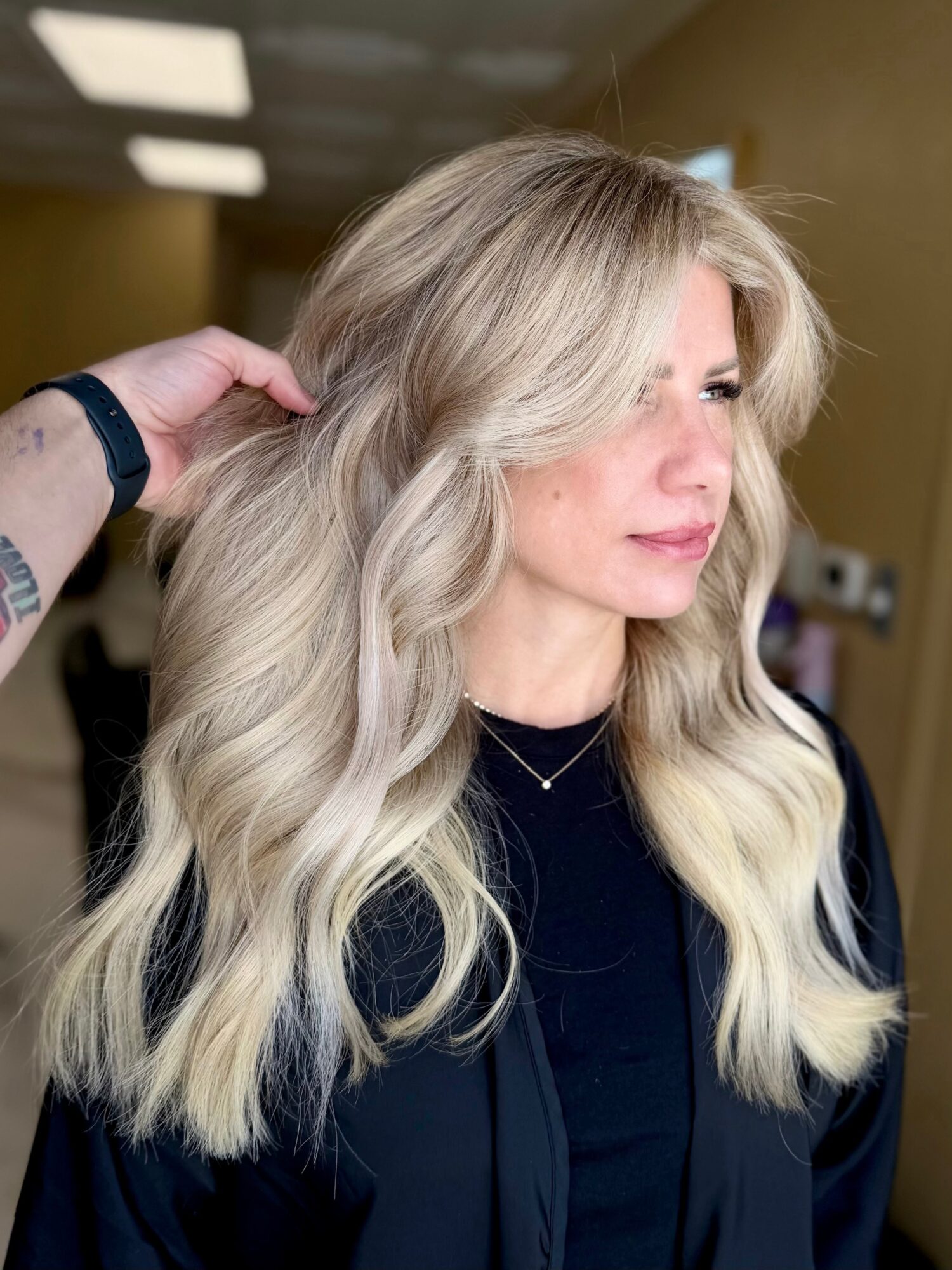 Woman with long, wavy blonde hair, wearing a black top, in a salon setting.