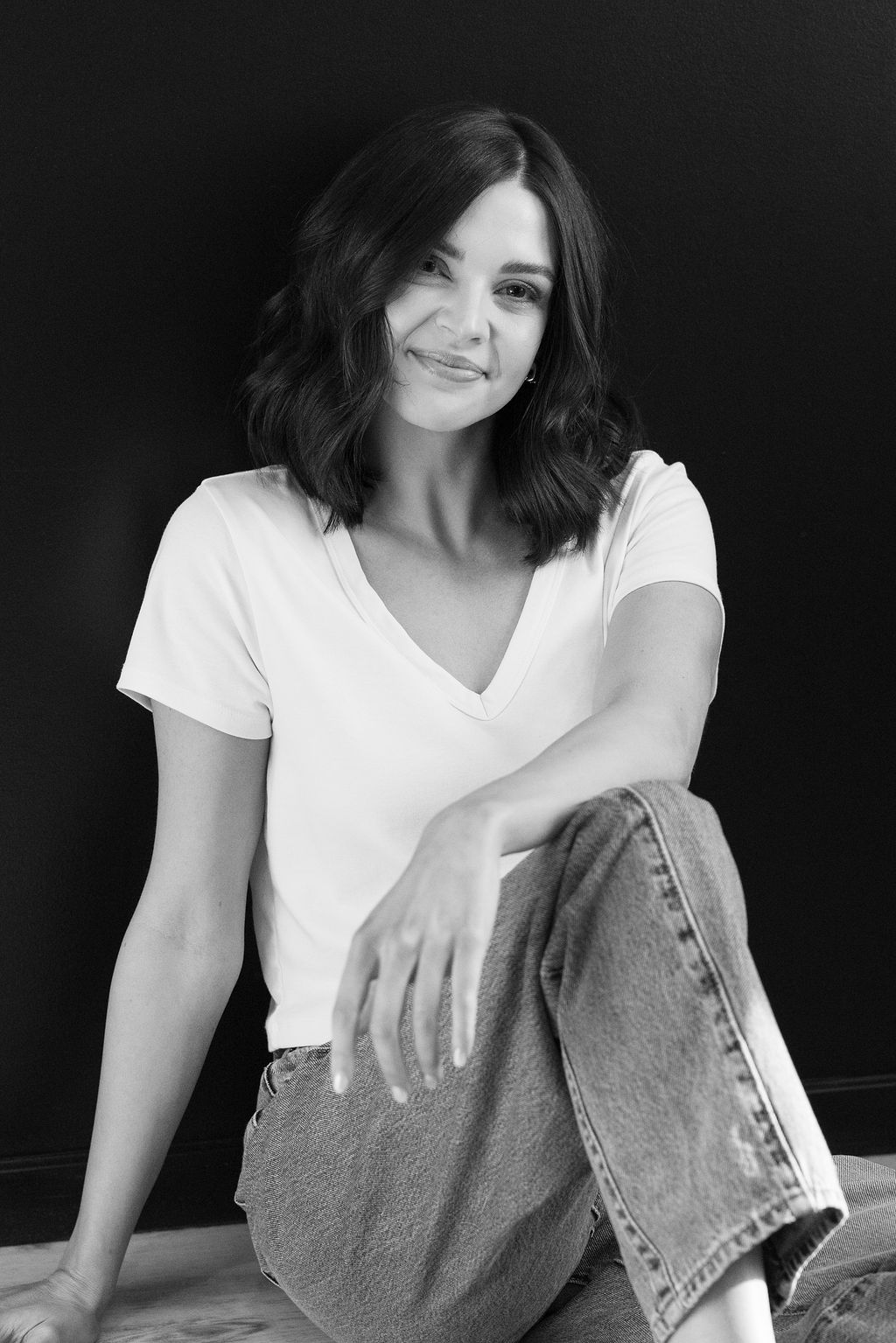 Woman with shoulder-length hair smiling, sitting with one knee up, wearing a white t-shirt and jeans, against a dark background.