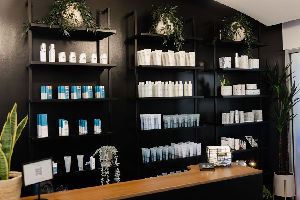 Black shelves filled with white and blue products, plants on top, wooden counter with small items, and potted plants nearby.