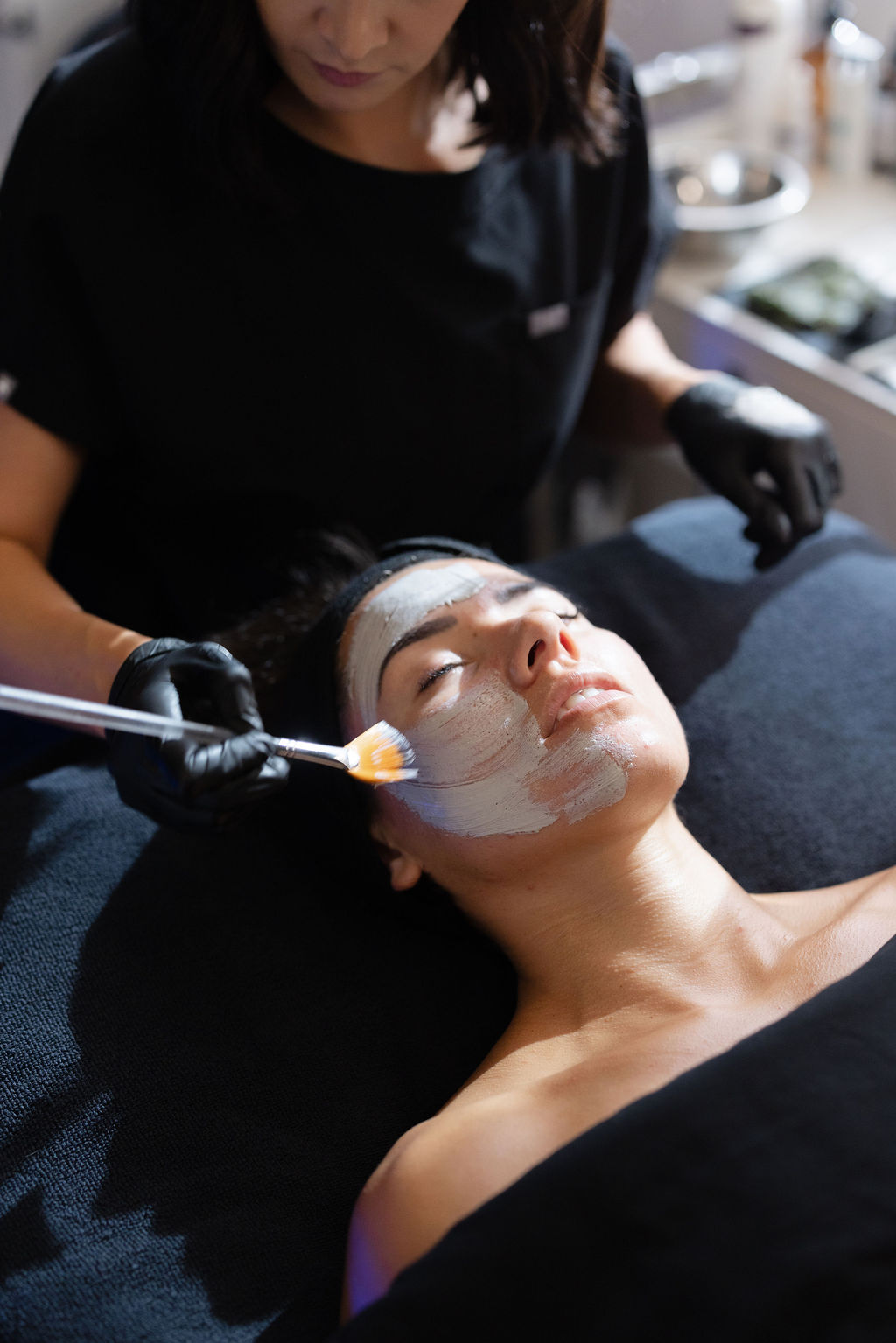 A woman receives a facial treatment with a mask applied, lying on a treatment bed, while a professional applies a product.